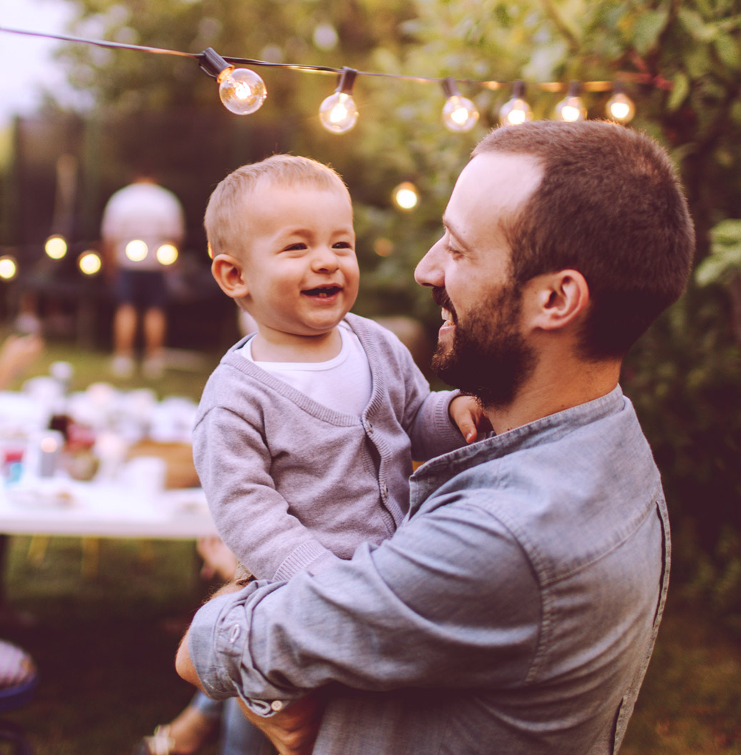 Man met baard met een lachende peuter in zijn armen in de tuin met op de achtergrond een lange tafel en sfeerverlichting 